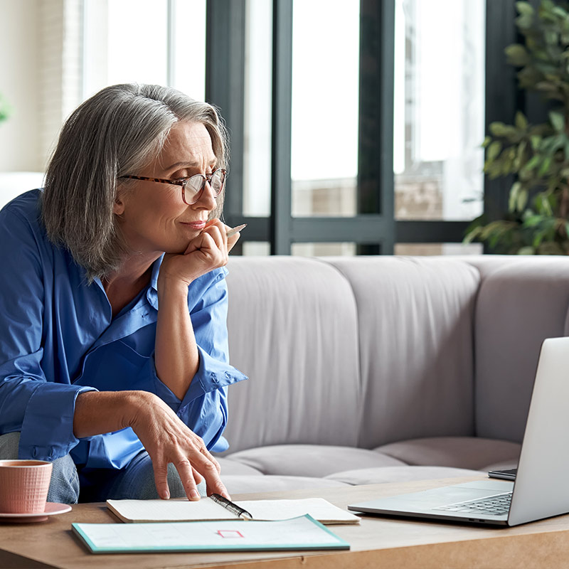 balance-thera Frau sitzt am Couch-Tisch, mit Block und Stift und schaut lächelnd in ihren Laptop